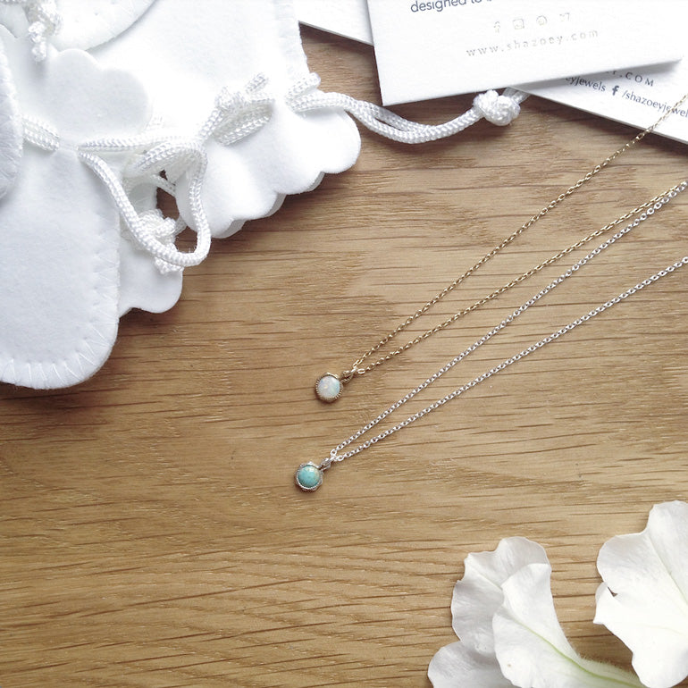 Two necklaces with small birthstone pendants on a wooden surface with white flowers.