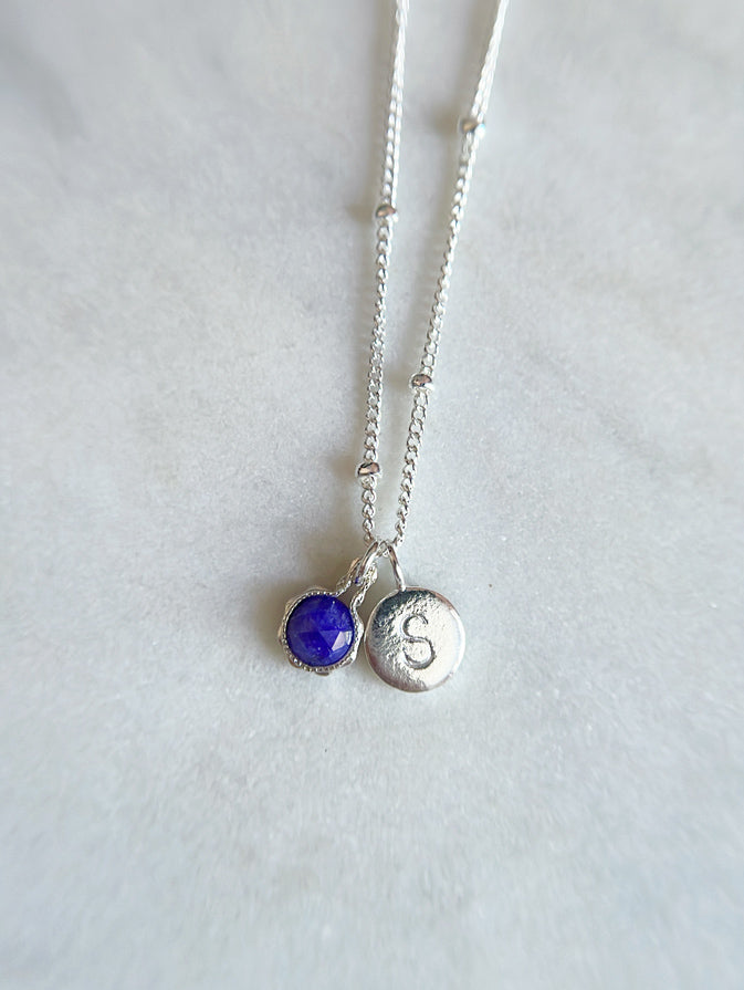 Sterling Silver necklace with a sapphire pendant and a round initial letter charm on a marble board