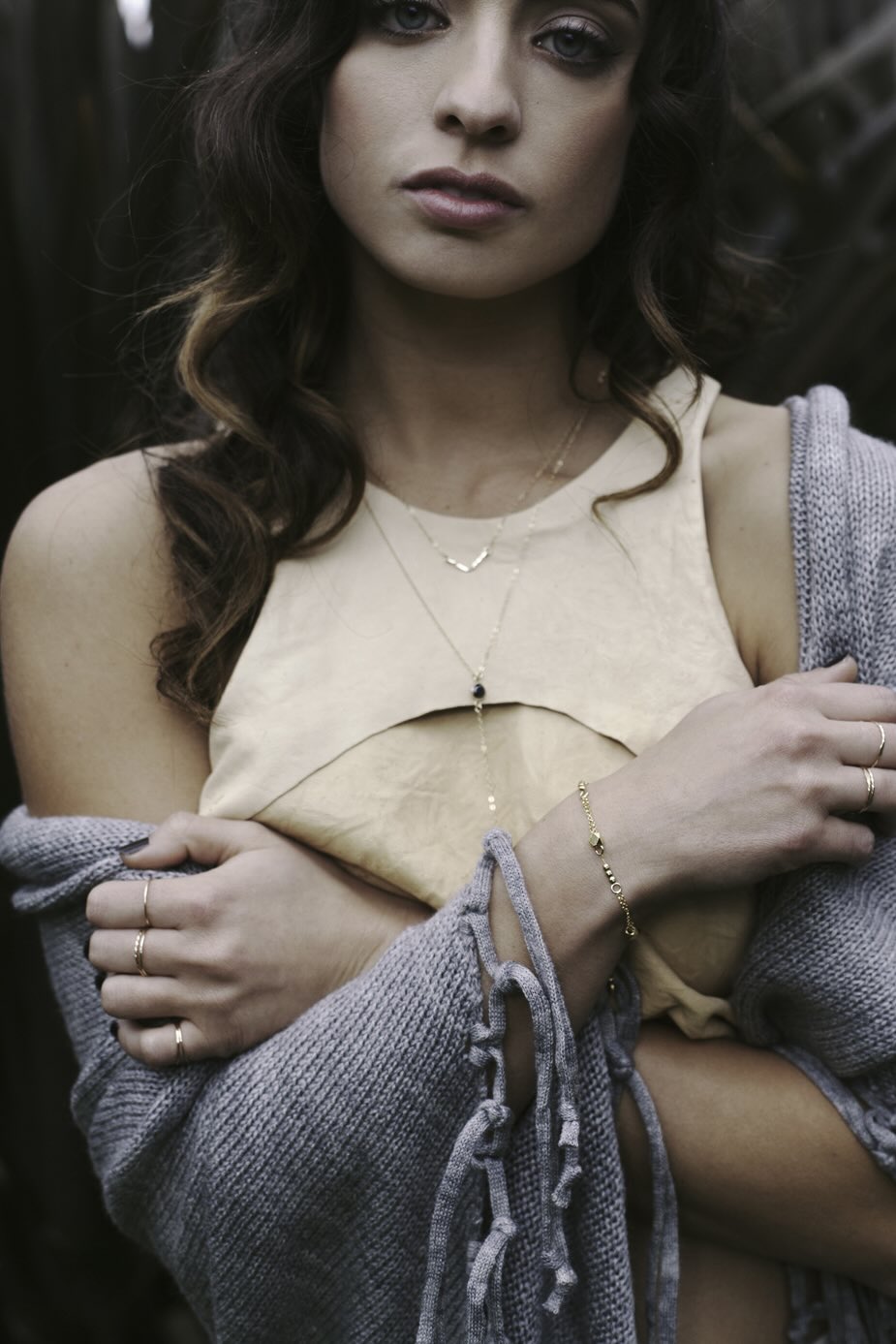 Woman wearing dainty layering gold necklaces and gold filled stacking rings, a beige top and grey cardigan.