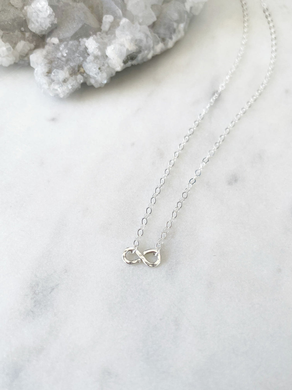 Dainty Silver infinity necklace on a marble surface with crystal rock in the background