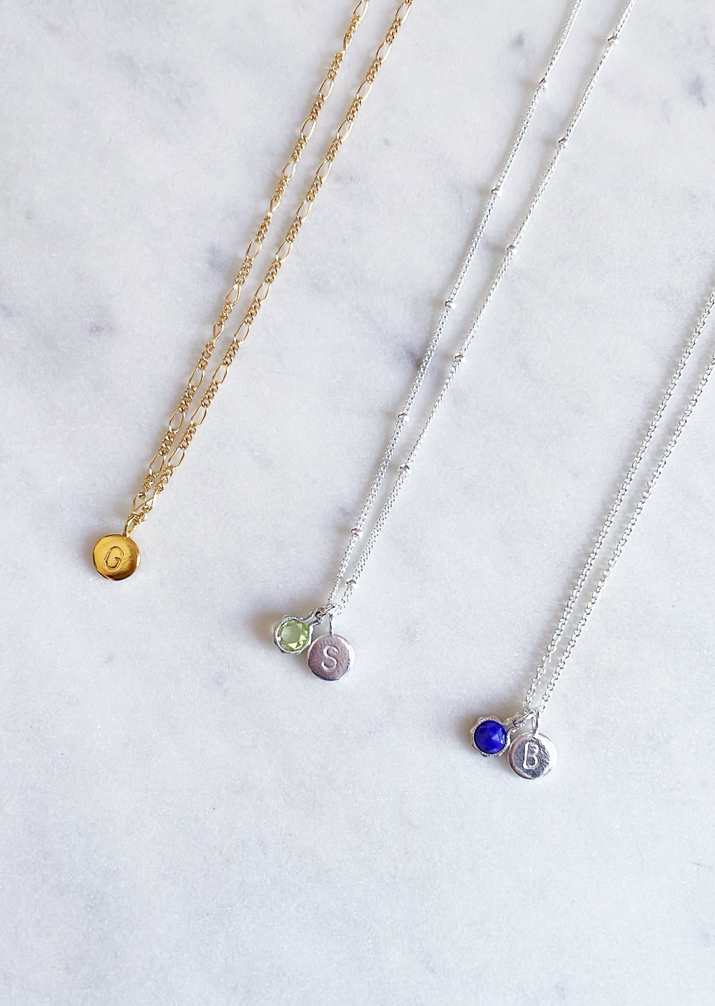 Sterling silver birthstone necklaces with peridot and blue sapphire gemstones and a gold initial necklace with figaro chain on a marble board